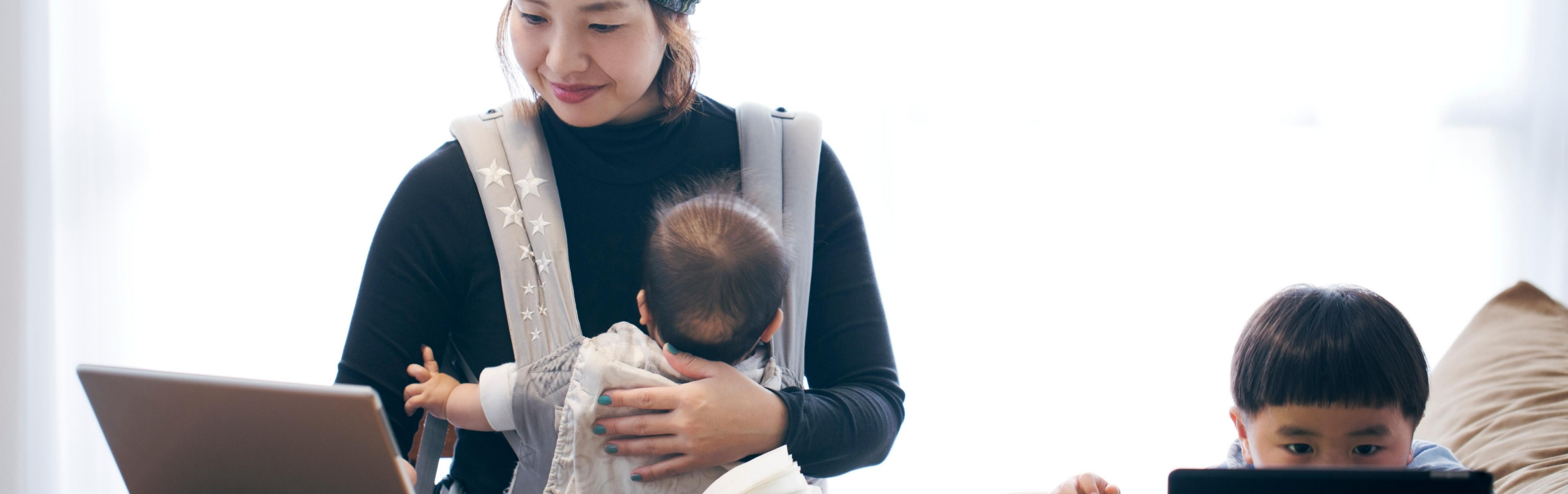 Mother works on her laptop at a table holding baby in baby carrier while her toddler sits next to her using tablet device