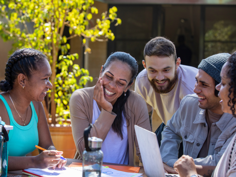 A group of First Nations students are happily interacting around a laptop at an outside table