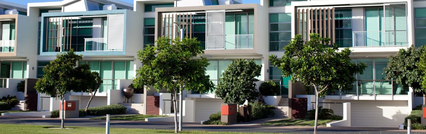 A view from the street of a row of modern double-storey multi-dwelling units with young trees planted along the nature strip