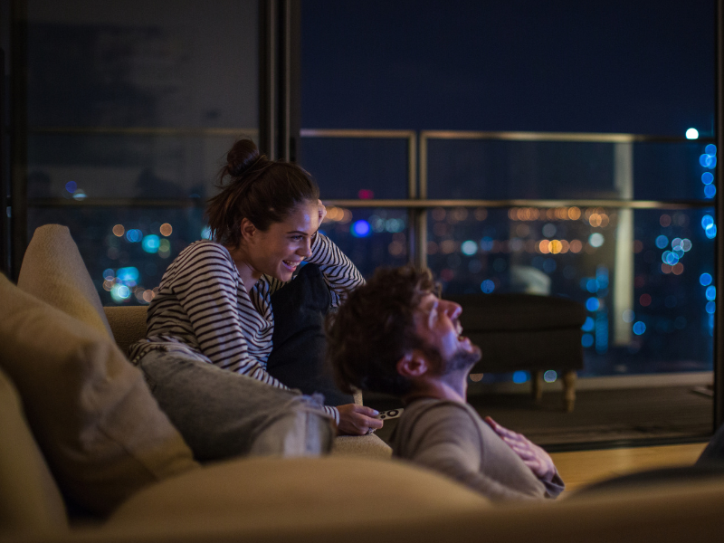 A happy couple are in the lounge room of their apartment at night watching the TV and laughing