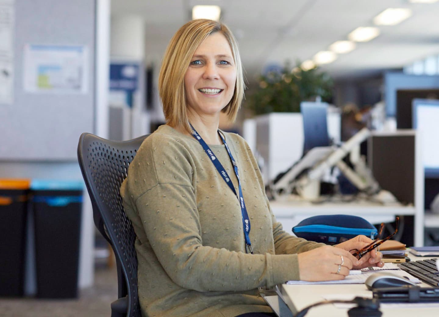 An nbn employee is sitting at a desk in the office working on a computer and is smiling at the camera
