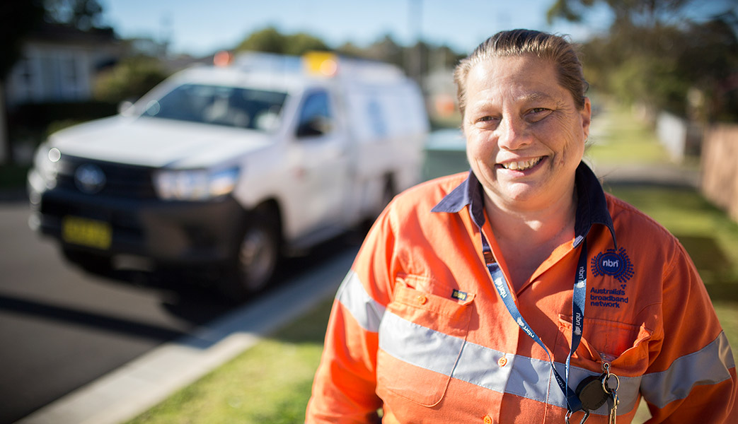 A smiling nbn field team member, Melanie, stands in a suburban street with her vehicle parked in the background